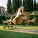 Monumental Classical Rearing Herd of Seven Horses Bronze Sculpture in polished gold by Giant Sculptures, displayed on lawn beside a stone bench near a villa.