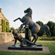 Monumental Classical Rearing Horse and Nude Male Figure Bronze Sculpture by Giant Sculptures, dark patina, displayed on a stone terrace beside a formal garden.