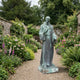 Monumental Classical Robed Saint Bronze Statue by Giant Sculptures with aged verdigris patina, standing on a stone plinth along a gravel path in a walled flower garden.