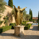 Giant Sculptures Monumental Classical Sacred Heart Jesus Bronze Statue in gilded gold, arms outstretched on a stone pedestal in a formal garden by a reflecting pool.