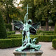 Monumental Classical Saint Michael Archangel Bronze Sculpture by Giant Sculptures with verdigris patina, raising a cross over a defeated figure in a formal garden.