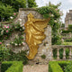 Monumental Classical Standing Angel Bronze Sculpture in gold finish by Giant Sculptures, wings spread, mounted on a stone garden wall amid climbing roses.