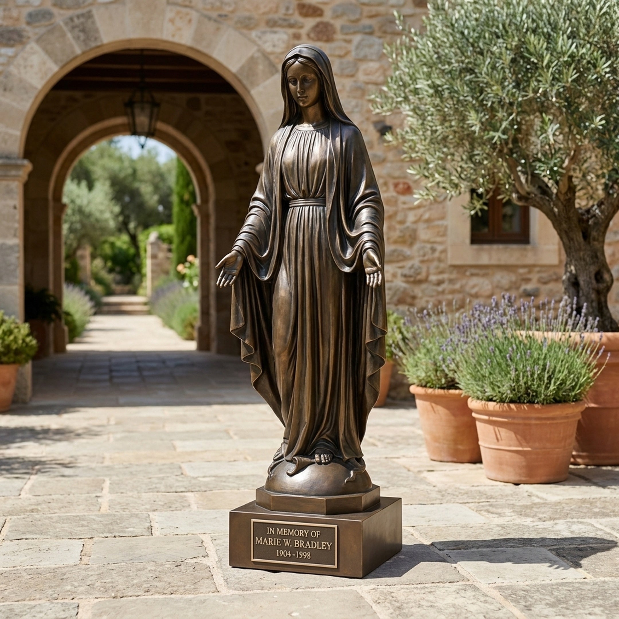 Monumental Classical Standing Virgin Mary Bronze Statue by Giant Sculptures on an engraved memorial plinth in a stone courtyard with olive tree and lavender.