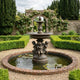 Giant Sculptures Monumental Classical Three Cherubs Fountain in golden-bronze with verdigris patina, set in a brick-edged lily pond within a formal hedged garden.