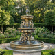 Three-tier bronze fountain by Giant Sculptures with seated draped figures, rearing horses, and a crowned figure on top, set in a formal rose garden.