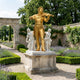 Monumental Classical Violinist and Allegorical Figures Bronze Sculpture in gold and white on a stone plinth, displayed in a formal lavender garden.