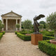 Monumental Classical Winged Goddess Bronze Sculpture by Giant Sculptures on a wooden plinth in a formal box hedge garden beside a columned stone pavilion.