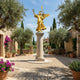 Giant Sculptures Monumental Classical Winged Victory Goddess Bronze Sculpture in gilt gold finish on a marble column, displayed in a Mediterranean courtyard with olive trees.