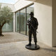 Monumental Contemporary Combat Soldier Bronze Statue by Giant Sculptures, standing on a circular base in a courtyard with olive tree and glass walls.