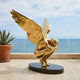 Monumental Contemporary Crouching Angel Bronze Sculpture in polished gold, wings sweeping upward, displayed on a dark oval plinth on an oceanview terrace.