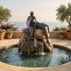Giant Sculptures Monumental Contemporary Dual Mermaid Bronze Fountain in warm patina, two figures on a boulder above a circular pool, with sea view beyond.