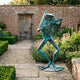 Monumental Flute-Playing Frog Bronze Sculpture by Giant Sculptures with verdigris patina, standing on a stone path beside lavender in a walled garden.