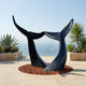 Monumental Interlocking Whale Tails Bronze Sculpture by Giant Sculptures, two black flukes arching from mulch on a coastal terrace with succulents and sea view.