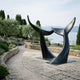 Monumental Interlocking Whale Tails bronze sculpture by Giant Sculptures, deep black patina flukes arching apart on a seaside terrace overlooking a harbour.