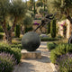 Monumental Contemporary Male Runner Bronze Sculpture by Giant Sculptures balanced atop a sphere on a stone plinth in a Mediterranean garden with olive trees.
