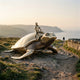 Monumental Man Riding Sea Turtle Bronze Sculpture by Giant Sculptures, polished gold shell and darker underside, displayed on a coastal cliff path at dusk.