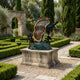 Monumental Contemporary Melting Clock and Figures Bronze Sculpture by Giant Sculptures on a stone plinth in a manicured hedge garden with cypress trees.