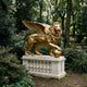 Gold-finished Monumental Heraldic Winged Lion Bronze Sculpture by Giant Sculptures, paw on globe atop a stone balustrade plinth in a wooded garden setting.