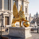 Monumental Heraldic Winged Lion Bronze Sculpture by Giant Sculptures in gold finish, roaring with paw on a globe, on a marble plinth outside a classical building.