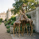Monumental Life-Size Giraffe Herd Bronze Sculpture by Giant Sculptures, three painted bronze giraffes clustered on a stone path beside a manor garden.