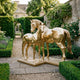Monumental Mare and Foal Bronze Sculpture by Giant Sculptures in polished gold finish, displayed on a gravel path in a manor garden with roses and hedges.