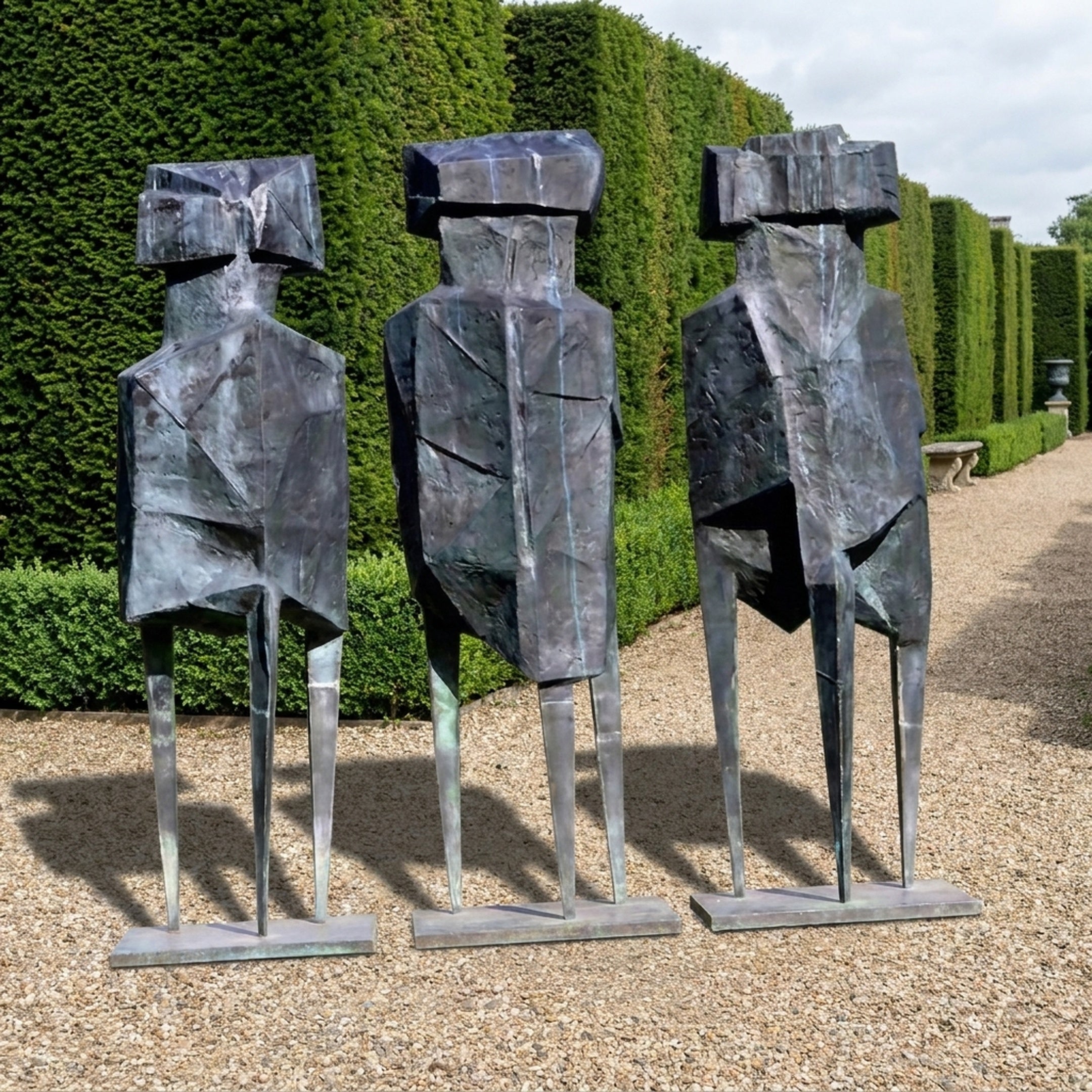 Monumental Trio of Abstract Geometric Figures bronze sculpture by Giant Sculptures, three angular faceted forms on stilted legs, displayed on gravel beside a hedge.