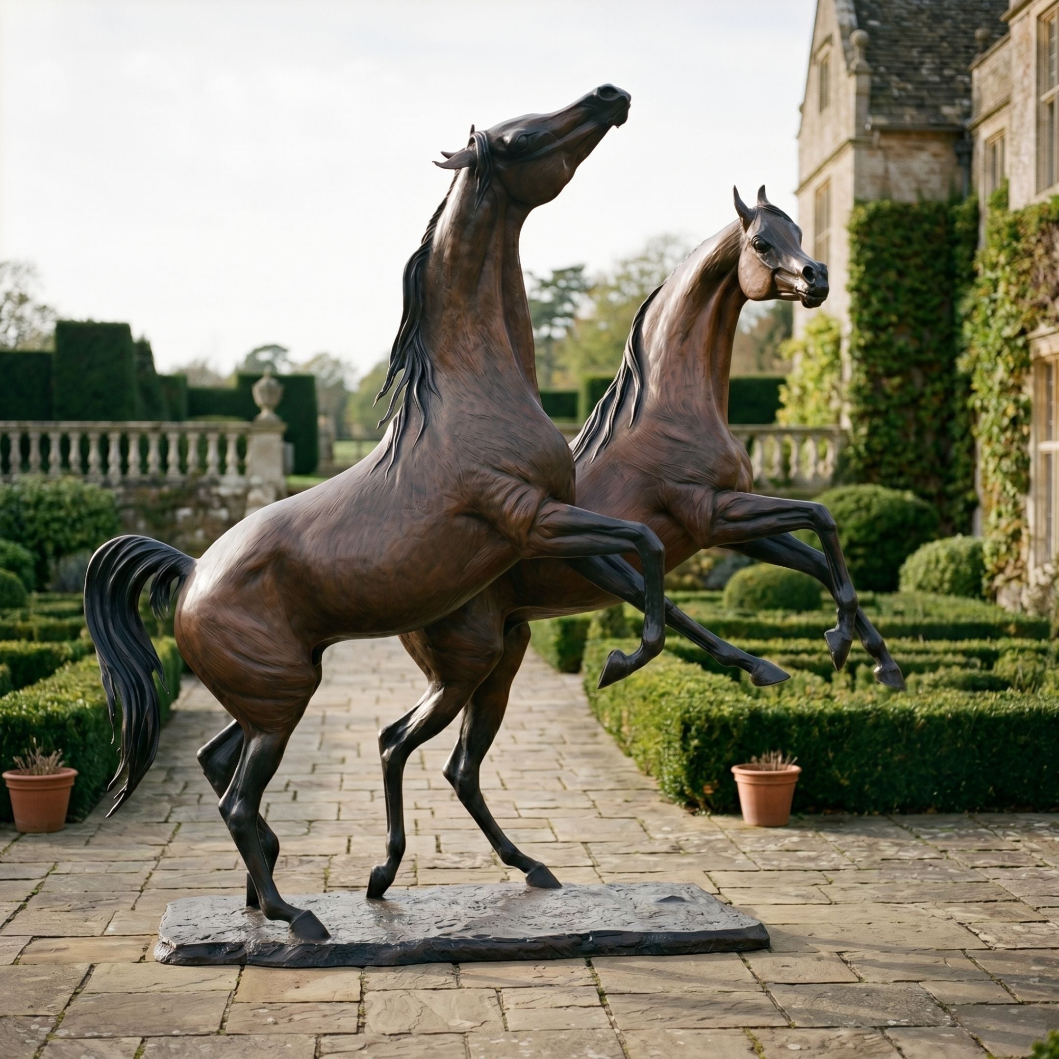 Monumental Rearing and Leaping Arabian Horses Bronze Sculpture by Giant Sculptures, warm brown patina, displayed on a stone garden path beside a manor.