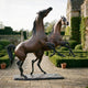 Monumental Rearing and Leaping Arabian Horses Bronze Sculpture by Giant Sculptures, warm brown patina, displayed on a stone garden path beside a manor.