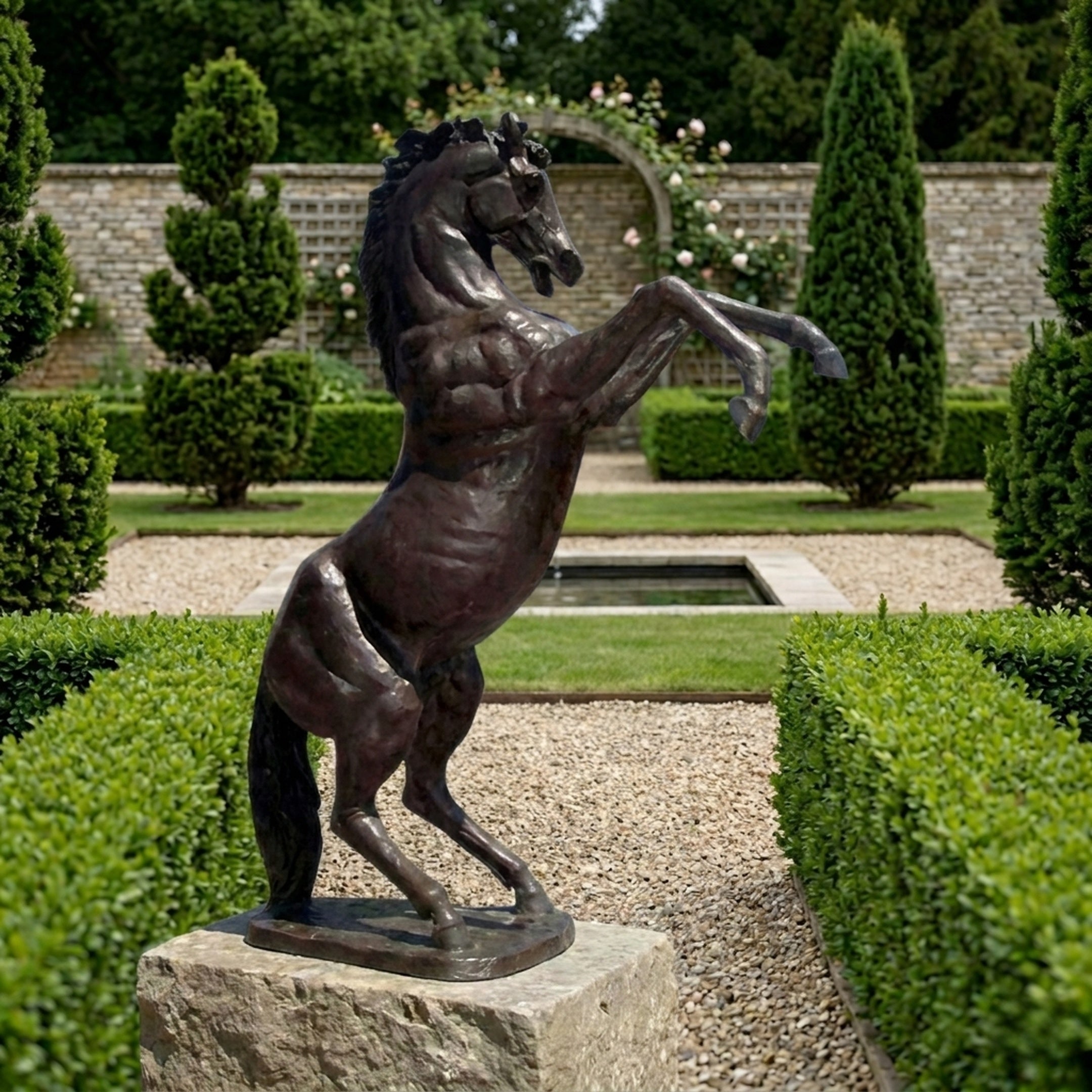 Monumental Rearing Horse Bronze Sculpture by Giant Sculptures with dark brown patina, mounted on a stone plinth in a formal garden with hedges and topiary.