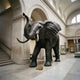 Giant Sculptures' 330cm Monumental Traditional African Elephant Bronze Sculpture displayed in a museum hall, trunk raised, ears flared, lighter-patina tusks.