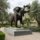 Monumental Traditional Bull Elephant Bronze Sculpture by Giant Sculptures with trunk raised and pale tusks, on a tiered stone plinth in an olive garden.