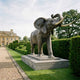 Monumental Traditional Bull Elephant Bronze Sculpture by Giant Sculptures, trunk raised and pale tusks forward, on a two-tier stone plinth beside manor hedging.