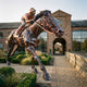 Monumental Traditional Racehorse and Jockey Bronze Sculpture by Giant Sculptures in copper-toned bronze, shown mid-gallop on a paved path beside a brick clock-tower building.