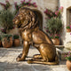 Monumental Traditional Seated Lion Bronze Sculpture by Giant Sculptures in warm golden patina, seated on a stone courtyard beside a fountain and pink blooms.