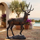 Monumental Traditional Walking Stag Bronze Sculpture by Giant Sculptures, 280cm, shown mid-stride on a rocky base in a sunlit Mediterranean poolside courtyard.