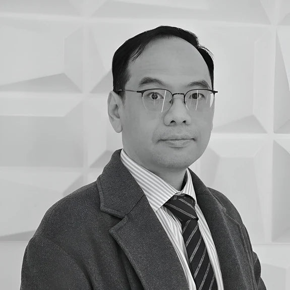 A person wearing glasses and a suit, including an overcoat and a striped tie, poses against a geometric patterned background. The image is in black and white.