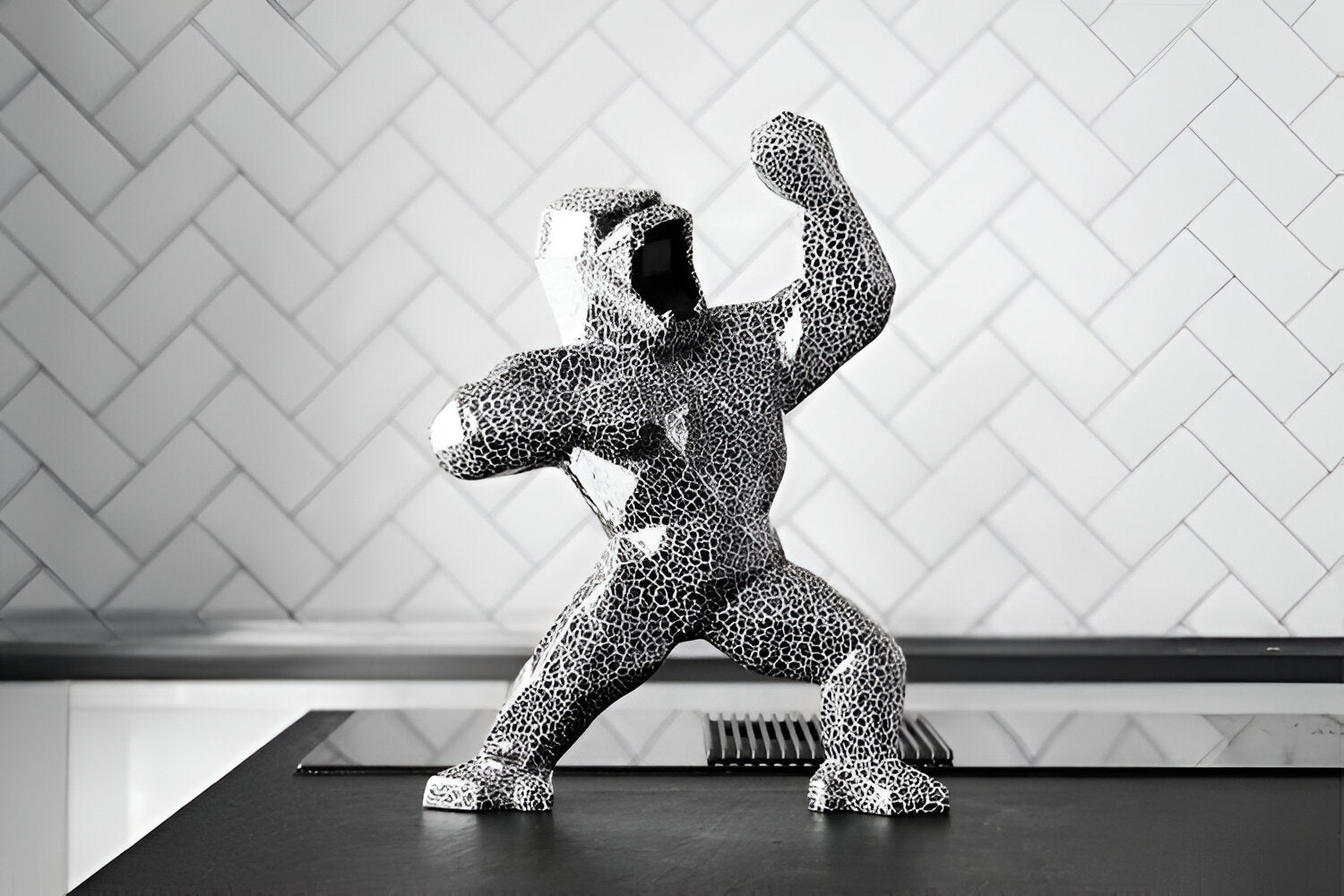 A black and white textured gorilla sculpture stands in a dynamic pose on a countertop. The background features a white chevron-patterned wall, adding a modern and stylish contrast to the striking figurine.