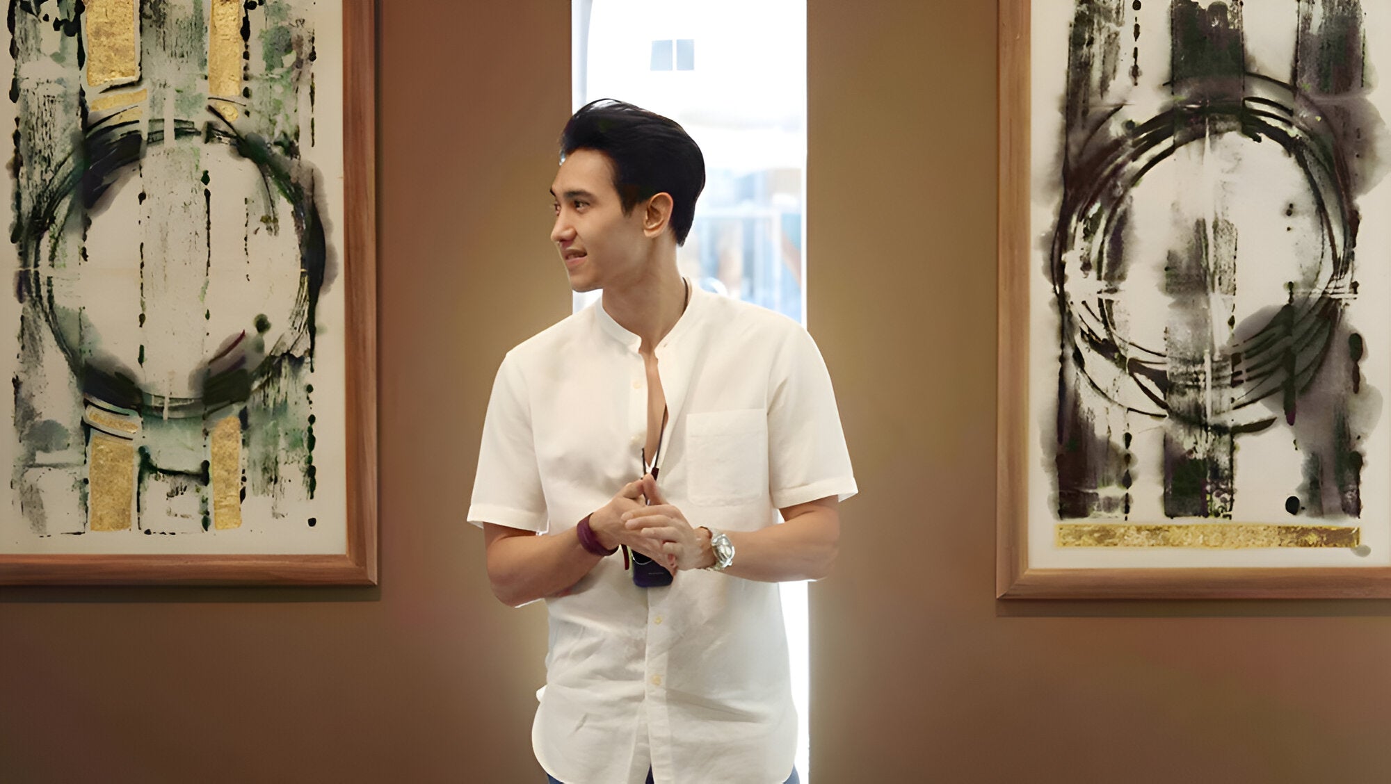 Man in a white shirt stands between two framed abstract paintings with circular patterns. He looks to his left, holding an object in his hand, in a room with brown walls and soft lighting.