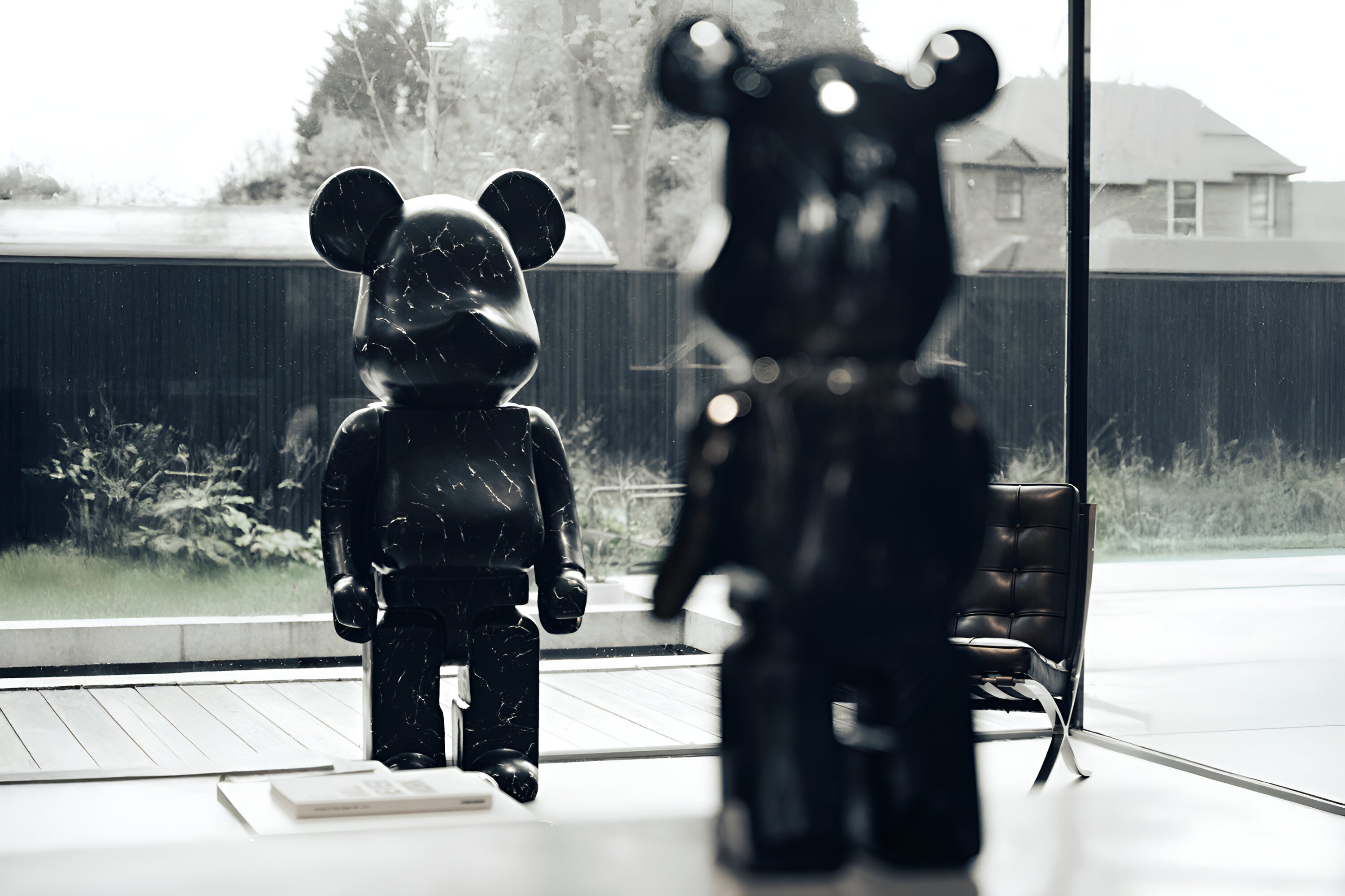 Two large, black bear-shaped sculptures with a marbled texture stand in a modern room. One is facing forward near a window overlooking greenery, and the other is closer to the camera, turned slightly sideways. A book rests nearby on a table.