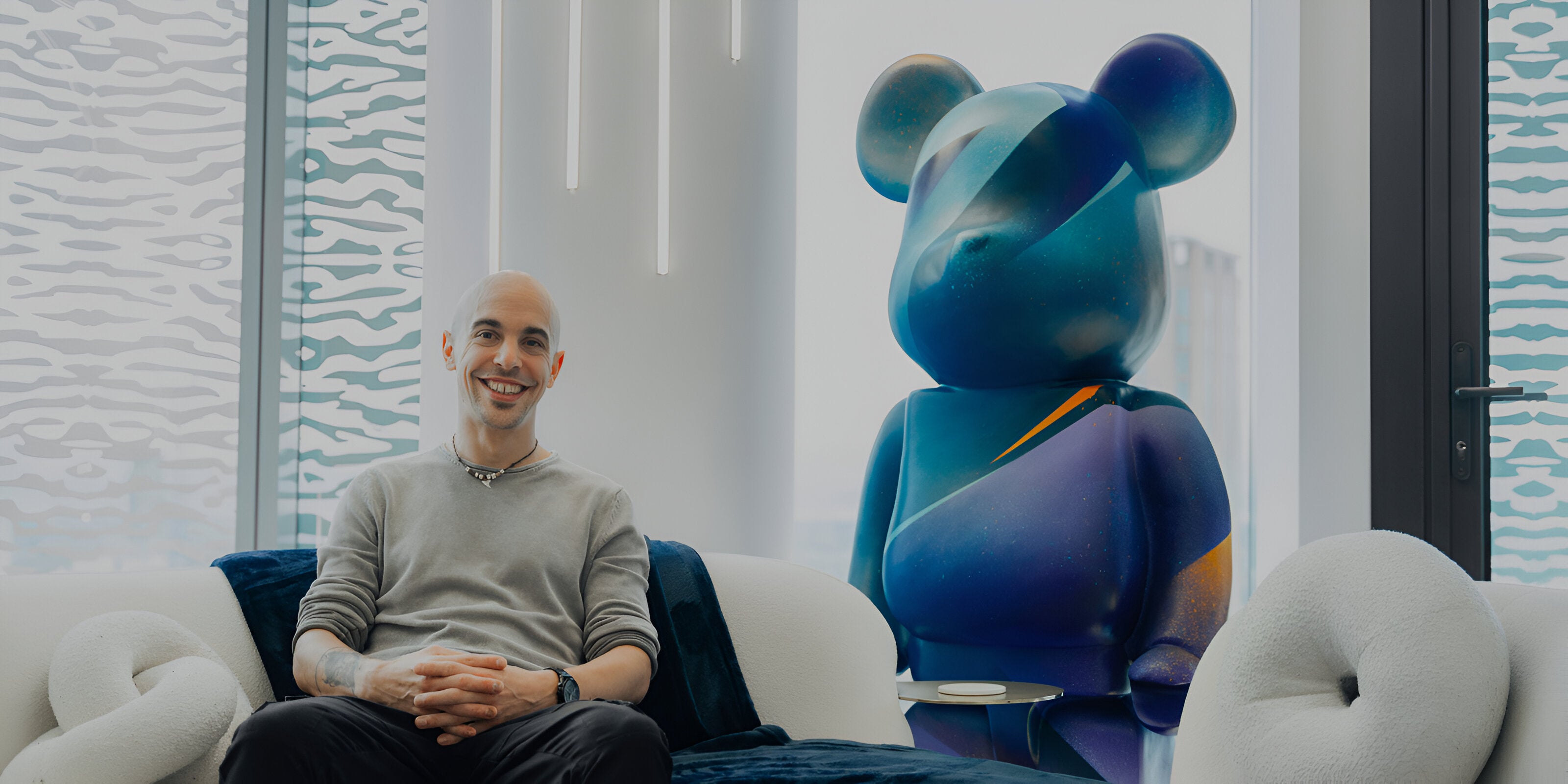 A person sitting on a modern, white sofa in a stylish room. Behind them is a large, colorful bear-shaped sculpture. The room has patterned glass windows and contemporary decor.