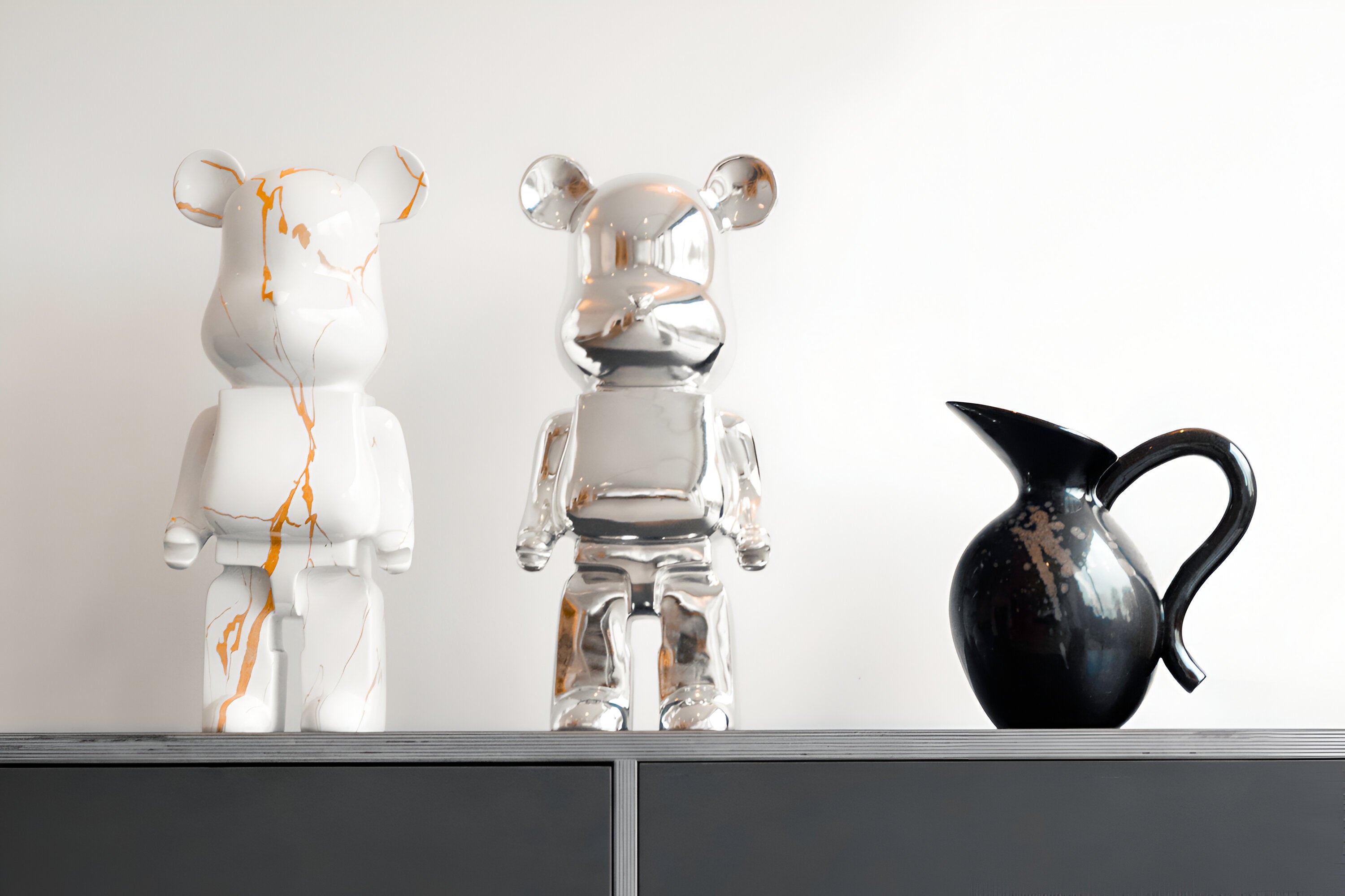 A pair of bear sculptures in a contemporary interior. One bear has a white marble finish with gold veining, while the other is a sleek, reflective silver. They are displayed on a black cabinet next to a glossy black vase, set against a white background.