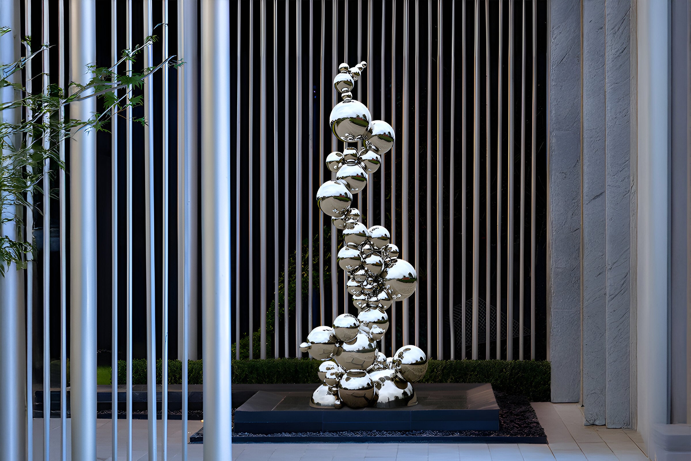 A modern outdoor sculpture composed of reflective, spherical shapes stacked vertically. It is set against a backdrop of vertical metallic rods, with greenery at the base. The sculpture exhibits a sleek, polished finish.