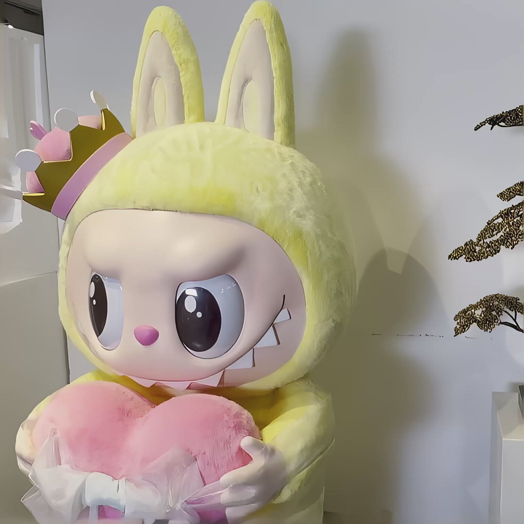 A video showcasing The Monster Let's Checkmate Heart Labubu Sculpture - 160cm, featuring a playful character in a yellow bunny suit with a crown, holding a large pink heart wrapped with a light blue bow.