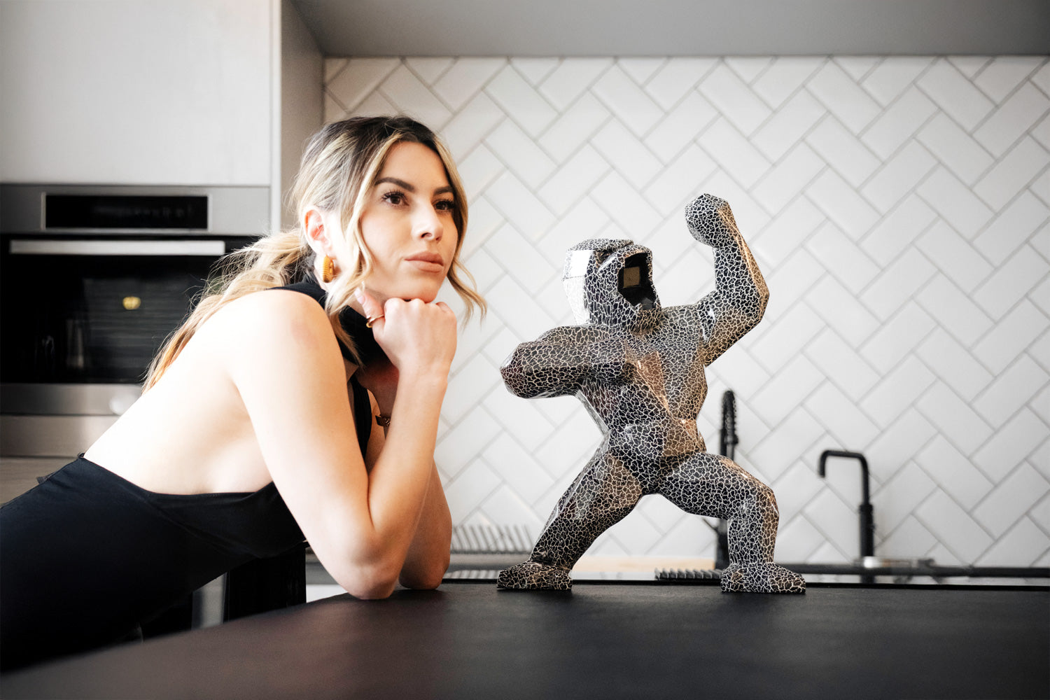 A person with long hair, wearing a black outfit, leans on a kitchen counter, gazing at a decorative sculpture of a flexing figure. The kitchen has white herringbone tile walls and modern appliances.