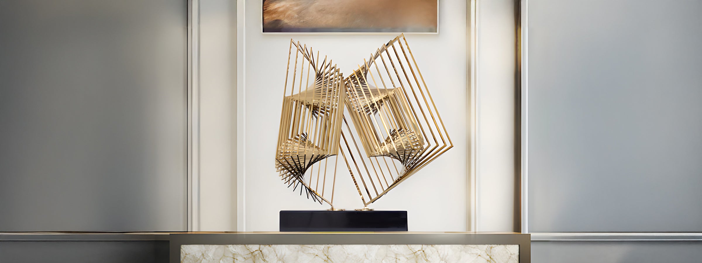 A modern, abstract sculpture featuring intersecting, geometric, gold rods displayed on a marble pedestal with a gold base. It is set against a backdrop of frosted glass panels and a framed artwork above.