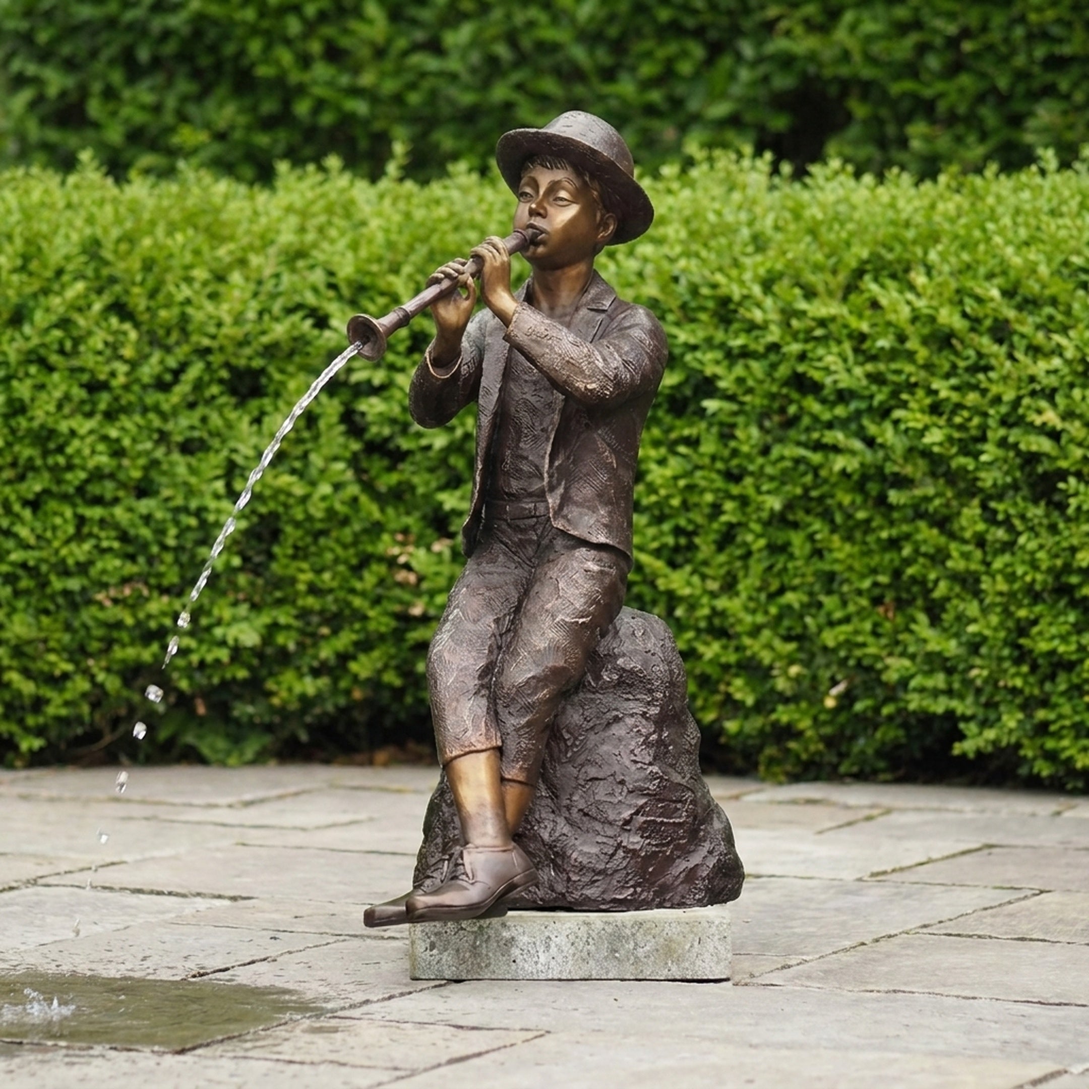 Traditional Boy Playing Flute Fountain Bronze Sculpture by Giant Sculptures, seated on a rock with water arcing from the flute, set on a garden patio.