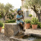 Traditional Flute Boy Bronze Fountain by Giant Sculptures on a stone block beside a pond, water jetting from the flute in a lavender-filled Mediterranean garden.