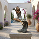 Traditional Girl with Shell Fountain Bronze Sculpture by Giant Sculptures, 110cm, seated on a wave-form base pouring water from a shell, displayed on a stone plinth in a sunlit Mediterranean courtyard.