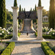 Traditional Liberty Bell Bronze Sculpture by Giant Sculptures, 91cm, suspended from a wooden yoke between two stone pillars in a formal garden with cypress trees.