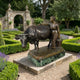 Traditional Milkmaid and Bull Bronze Sculpture by Giant Sculptures on a green marble plinth, displayed in a formal hedged garden with stone archway behind.